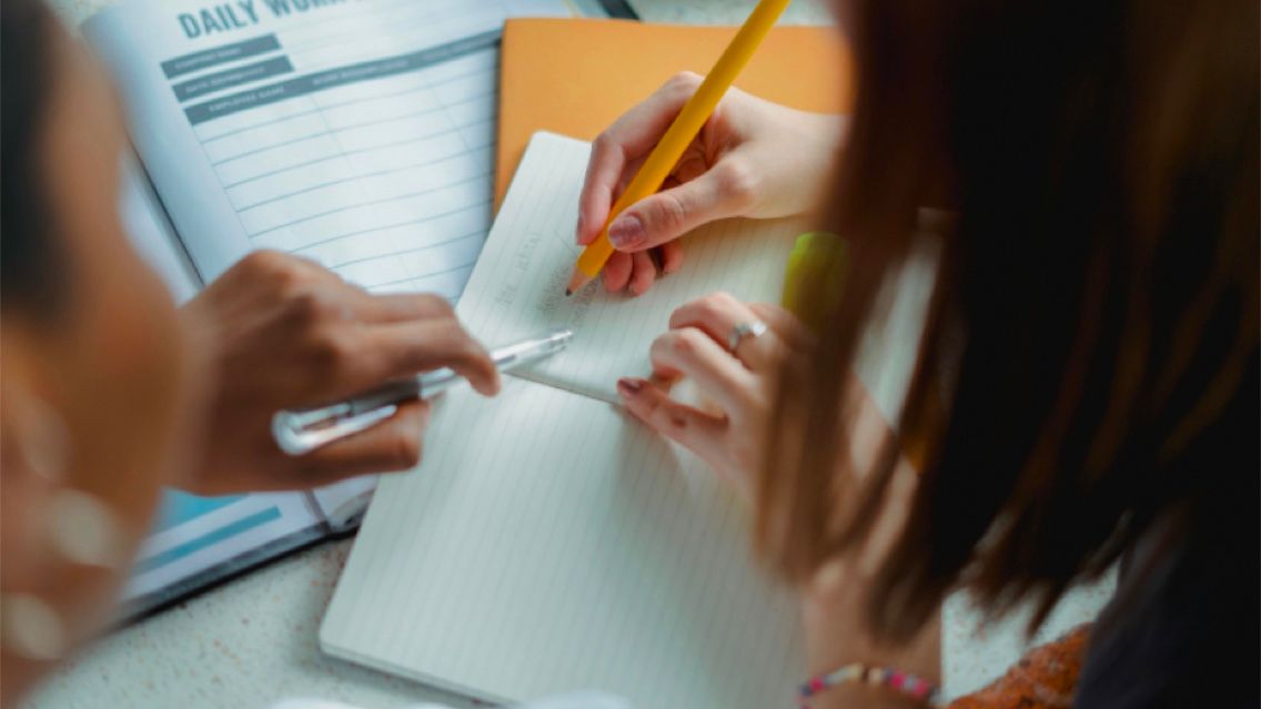 Two people look over a pile of papers while the one on the right takes notes